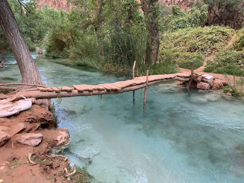 A wooden bridge spans a clear, turquoise stream surrounded by lush greenery and rocky banks.