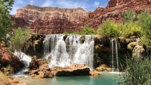 A scenic waterfall cascades into a turquoise pool, surrounded by lush greenery and red rock formations.