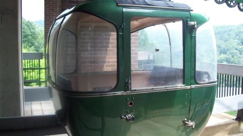 A green gondola cabin with large windows, set against a backdrop of trees and hills.
