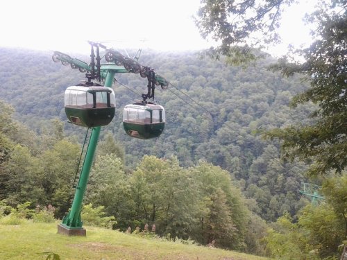 Two green gondolas on a cable car system, surrounded by lush green mountains and trees in the background.