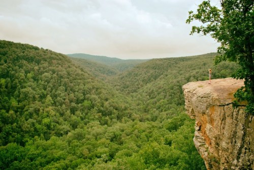 A person stands on the edge of a rocky cliff, overlooking a lush green valley surrounded by mountains.