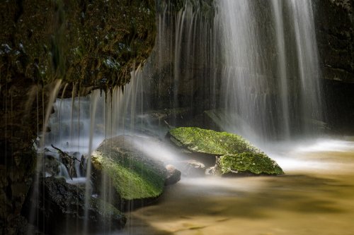 A serene waterfall cascades over moss-covered rocks into a calm, muddy pool below.
