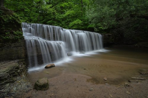 A serene waterfall cascades over rocks into a calm pool, surrounded by lush green trees and foliage.