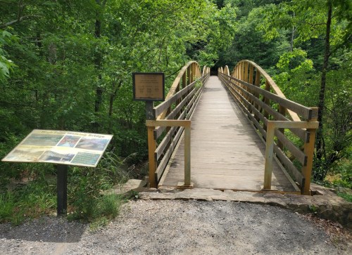 A wooden bridge arches over a path, surrounded by lush greenery, with an informational sign nearby.