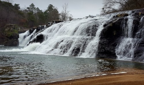 A serene waterfall cascading over rocks into a calm pool, surrounded by trees and a cloudy sky.