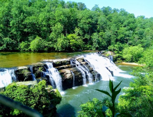 A serene waterfall cascades over rocky ledges, surrounded by lush green trees and a calm river.