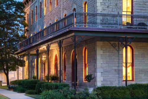 Historic stone building with a balcony, illuminated windows, and lush greenery in the foreground at dusk.