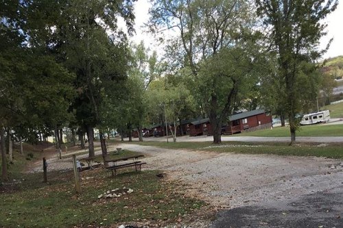 A gravel path leads through a wooded area with cabins in the background and a picnic table nearby.