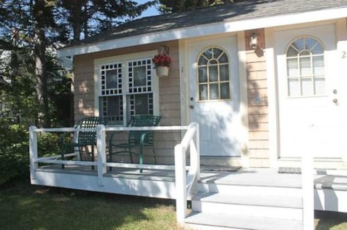 A cozy cottage with a small porch, two doors, and a window adorned with flowers. Green chairs are on the porch.