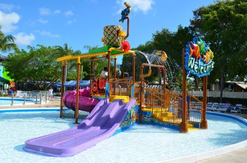 Colorful water play structure with slides and water features in a sunny pool area, surrounded by palm trees.