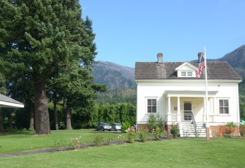 A white house with a porch and American flag, surrounded by green grass and trees, with mountains in the background.