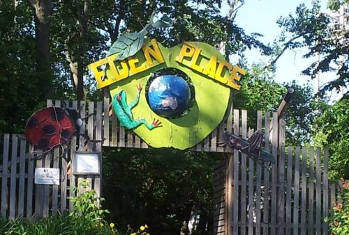 Colorful entrance sign to "Eden Place," featuring a globe, a frog, and insects, surrounded by lush greenery.