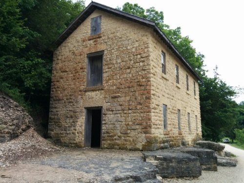A weathered stone building with boarded windows, surrounded by greenery and rocky terrain.