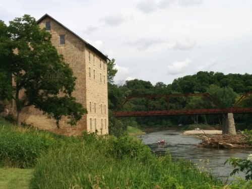 Historic stone building beside a river, with a red metal bridge in the background and lush greenery along the bank.