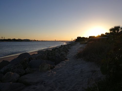 Sunset over a calm beach, with rocks lining the shore and silhouettes of trees in the background.