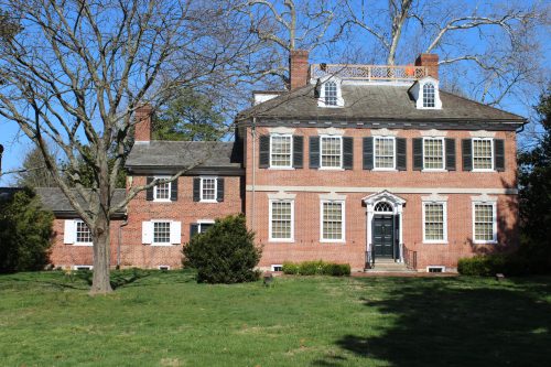 A large brick house with white trim, multiple windows, and a front porch, surrounded by a grassy lawn and trees.