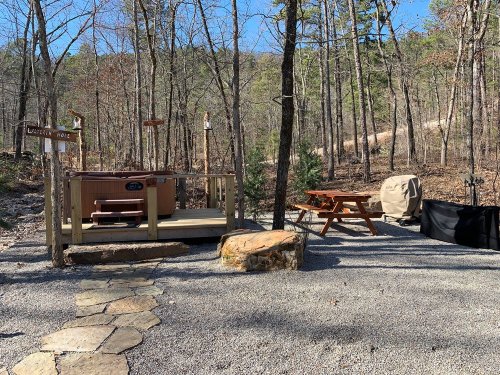 A cozy outdoor area with a hot tub, picnic table, and stone path, surrounded by trees and a clear blue sky.