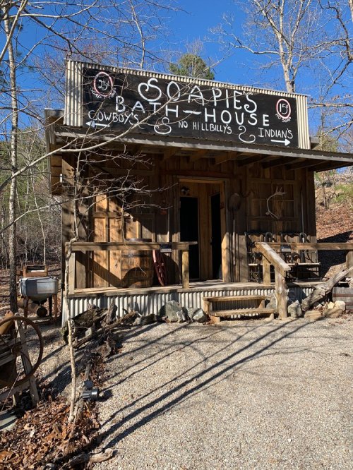 A rustic wooden bathhouse with a sign reading "Soapies Bath House" surrounded by trees and gravel.