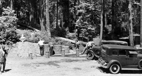 Historic black-and-white photo of people at a stone entrance in a forest, with vintage cars parked nearby.