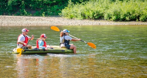 Three people kayaking on a calm river, surrounded by greenery, enjoying a sunny day outdoors.