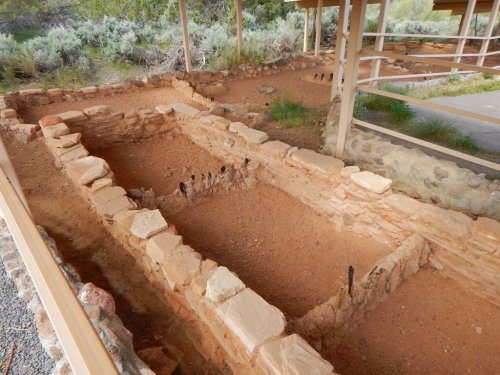 Archaeological site with stone foundations and a sandy floor, surrounded by a wooden structure and desert vegetation.