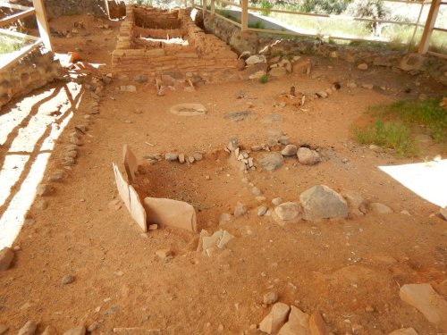 Archaeological site with stone foundations and circular features on sandy ground, surrounded by rocks and vegetation.
