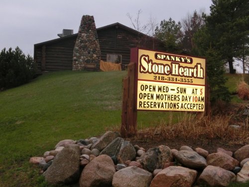 Sign for Spanky's Stone Hearth, with hours and reservation info, in front of a rustic log building and stone chimney.