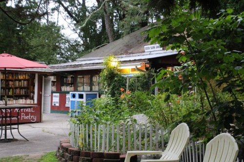 A cozy outdoor view of a small bookstore with a garden, featuring a patio and a sign for poetry events.