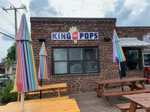 Exterior of a brick building with a sign reading "King of Pops" and colorful umbrellas over wooden picnic tables.