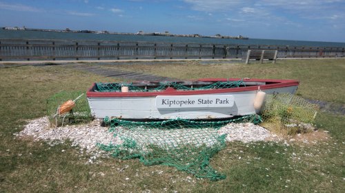 A weathered boat labeled "Kiptopeke State Park" surrounded by shells and fishing nets on a grassy area by the water.