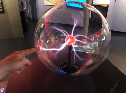 A person points at a plasma globe, with electric arcs radiating from a central red sphere inside a glass dome.