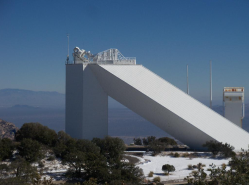 A large white structure resembling a ski jump, set against a clear blue sky and mountainous landscape.