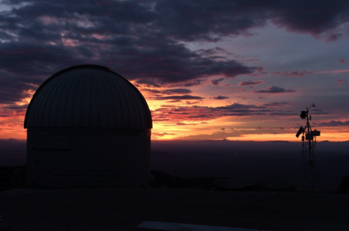 A silhouette of an observatory dome against a vibrant sunset with clouds and distant mountains.