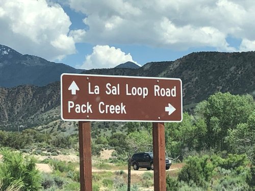 Brown road sign directing to La Sal Loop Road and Pack Creek, with mountains and greenery in the background.
