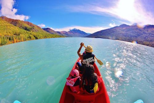 A group of children paddles a red canoe on a turquoise lake surrounded by mountains and vibrant autumn foliage.