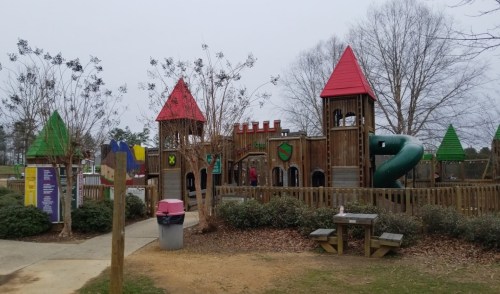 Colorful wooden playground resembling a castle, featuring slides, towers, and surrounding trees.