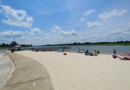A sandy beach by a lake with people relaxing, umbrellas, and a pier under a bright blue sky with fluffy clouds.