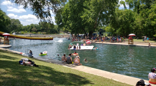 A busy park with people swimming and relaxing by a river, featuring a water slide and lush green trees.
