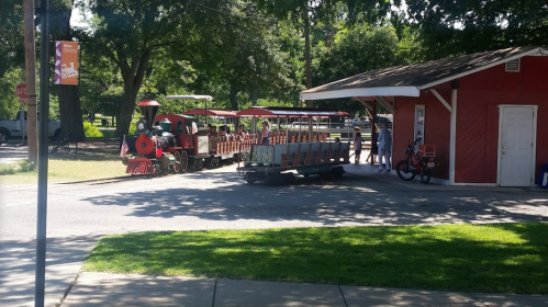 A red train with open cars is parked near a small red building in a park, surrounded by trees and grass.