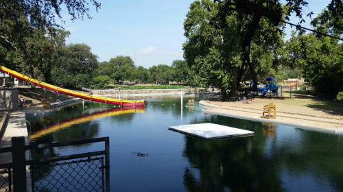 A serene park scene featuring a water slide, a swimming area, and playground equipment surrounded by trees.