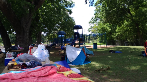 A park scene with a playground in the background, scattered blankets, and tents on the grass.