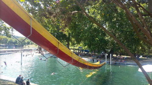 A colorful water slide extends over a swimming area with people enjoying the water and trees in the background.