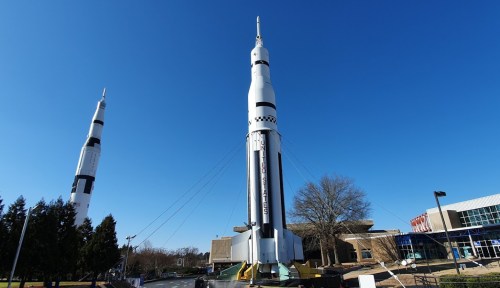 A large rocket stands tall against a clear blue sky, with trees and a building in the background.
