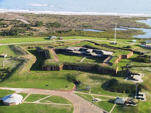 Aerial view of a historic fort surrounded by green fields and coastline, with buildings and pathways visible.