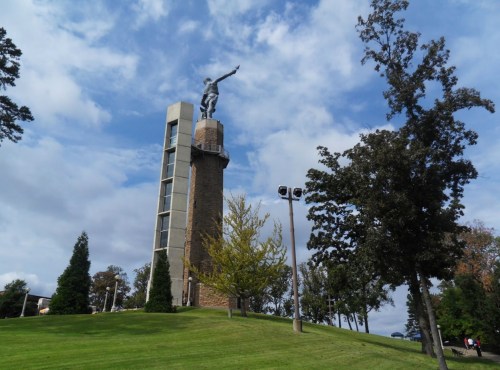 A tall statue atop a stone tower, surrounded by trees and a grassy area under a partly cloudy sky.