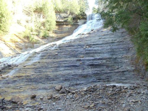 A cascading waterfall flows down a rocky slope, surrounded by trees and natural greenery.