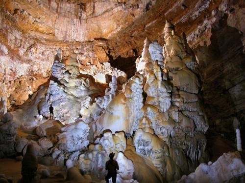 Two explorers in a cave surrounded by large, intricate stalagmites and stalactites illuminated by soft lighting.