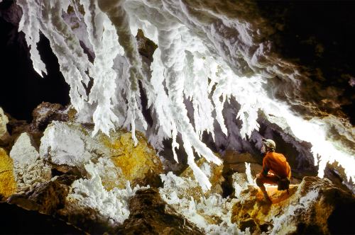 A person in an orange suit sits beneath stunning white stalactites in a cave, illuminated by soft light.