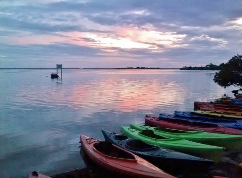 Colorful kayaks lined up on the shore at sunset, reflecting hues in the calm water under a cloudy sky.