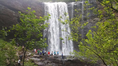 A group of people stands near a large waterfall surrounded by lush greenery and rocky terrain.